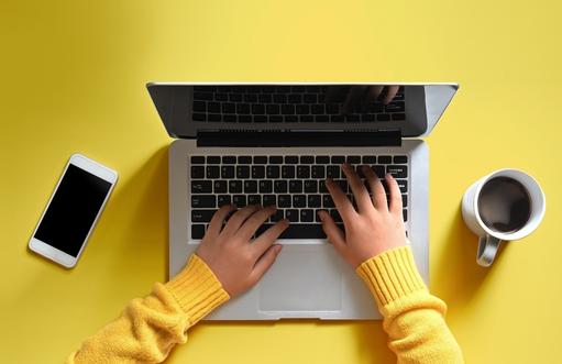 hands of female translator in yellow sweater typing on a laptop with coffee cup and phone nearby against a yellow background