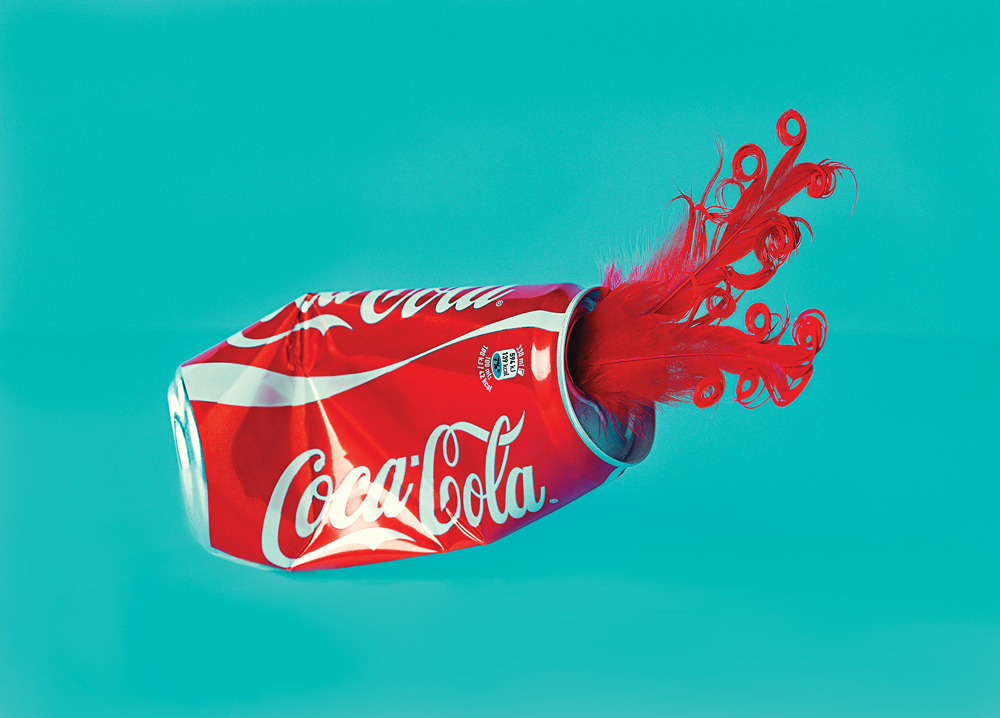 smashed coca-cola can with red feather coming out agains a turquoise backround
