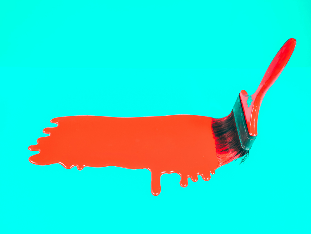 red brush applying red paint against a light blue background
