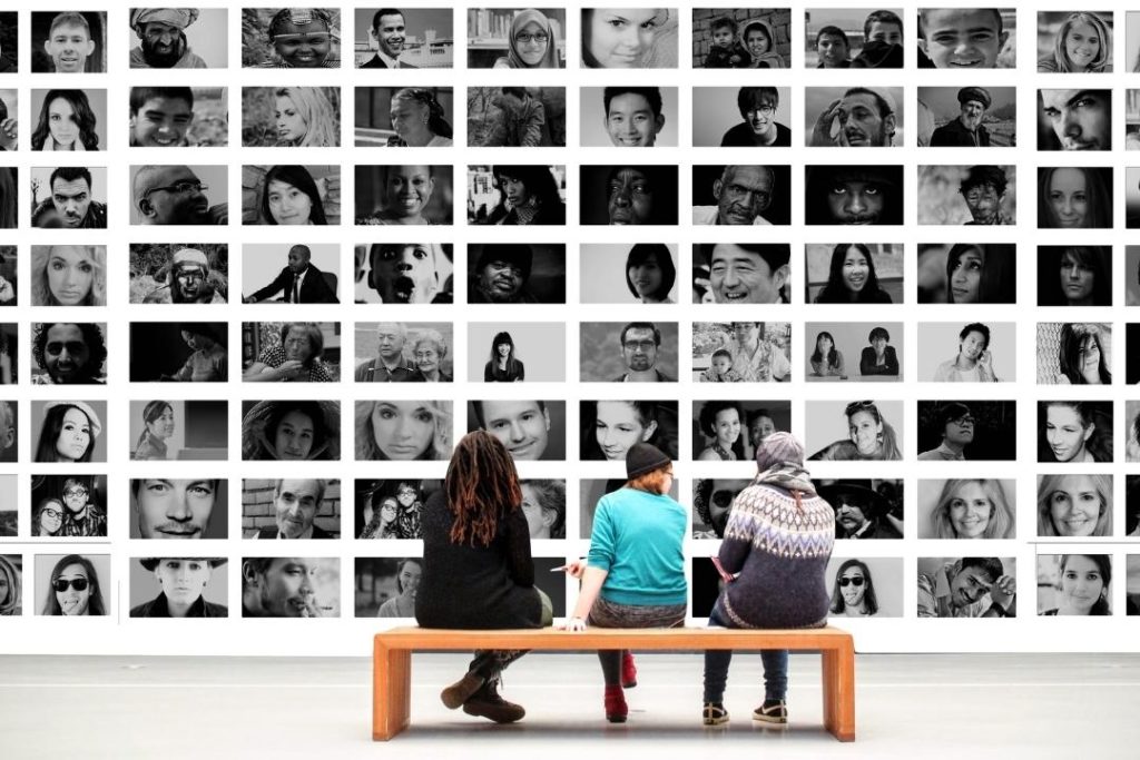 three people of different cultures sitting on a bench in gallery looking at a wall filled with black and white photos of people from all over the world