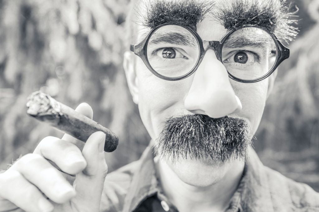 black and white headshot of man wearing clown glasses, nose and moustache and a cigar in his hand